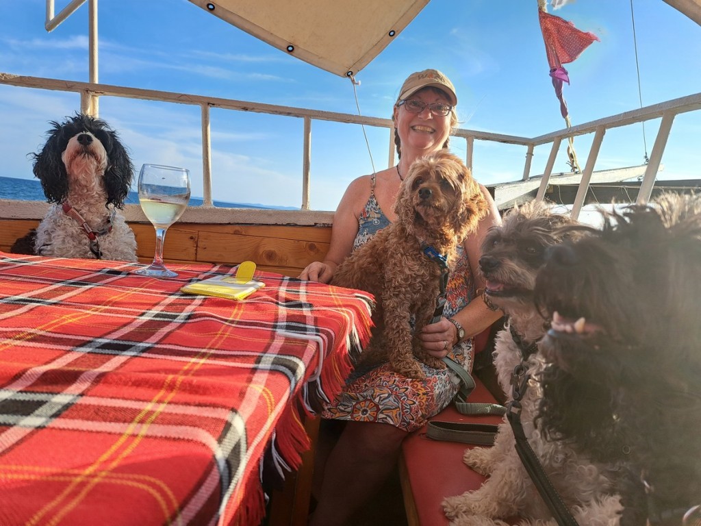 A photo of Jacqueline Lambert Author with The Fab Four, her cavapoos, (cavalier/poodle cross_ dogs. Jacqueline is wearing a baseball cap and is sitting at a dining table on a sunny day in the bow of the boat surrounded by the four dogs.