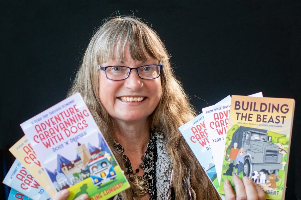 A headshot of author Jacqueline Lambert holding her seven published memoirs about overland travel with dogs including the award winning memoir Building The Beast
