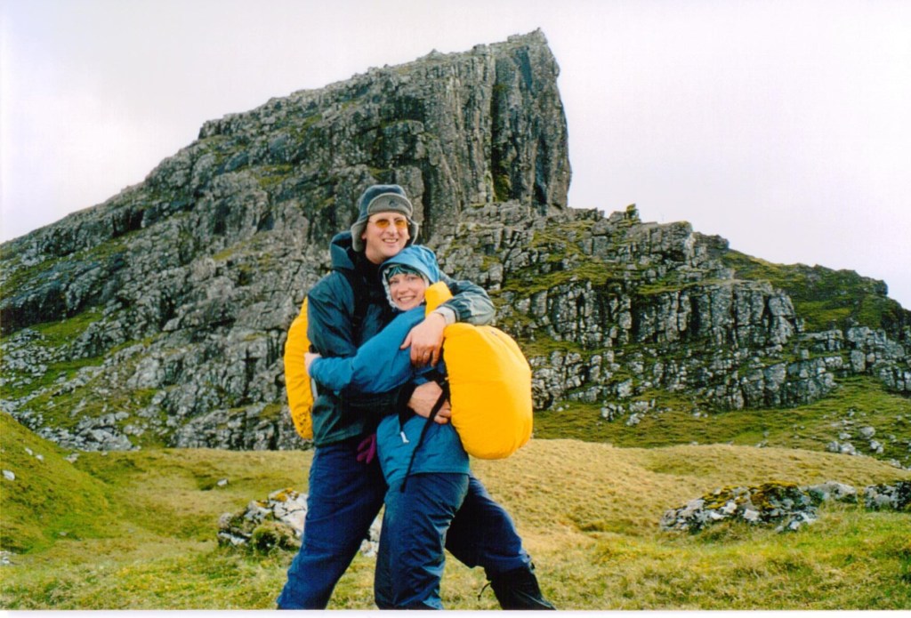 Author Jacqueline Lambert hugging husband Mark. Both are dressed for hill walking with backpacks, with a rocky mountain behind. 
