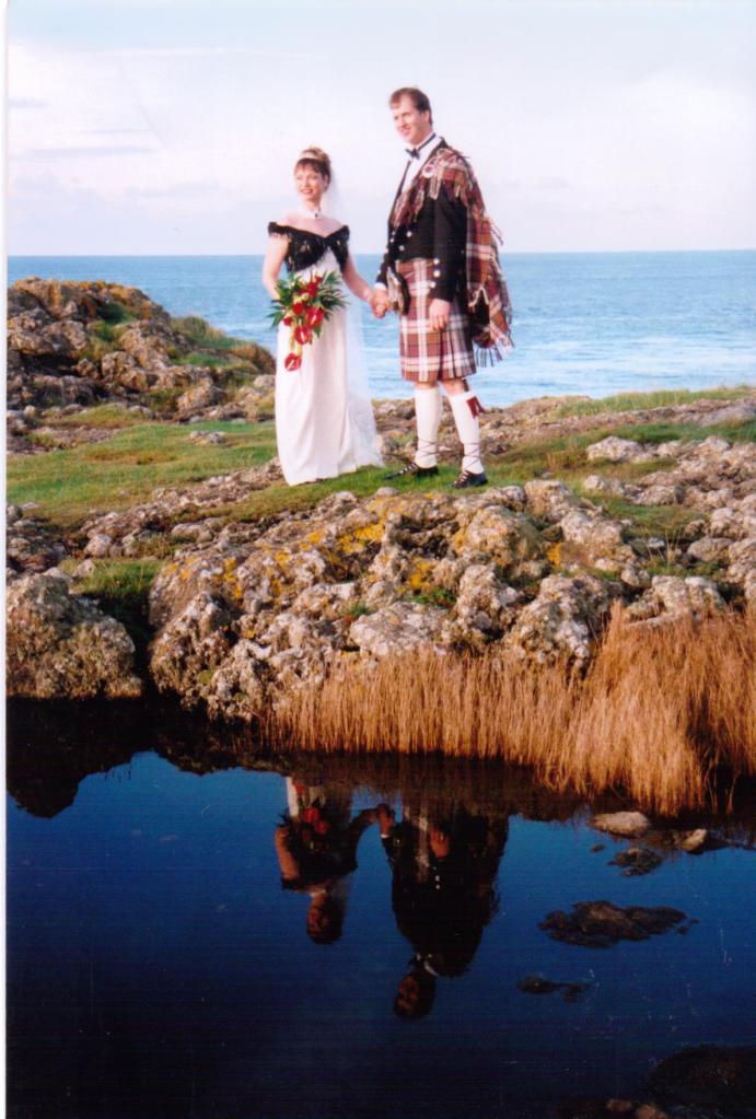 Author Jacqueline Lambert marrying husband Mark on the rocks outside a Scottish lighthouse. Mark is wearing a kilt and full Scottish garb, in dress McDonald tartan which is affiliated to clan Lambert The couple are reflected in a rock pool